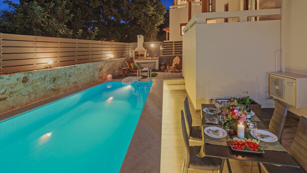 Ein Swimmingpool mit Holzterrasse, ein Essbereich mit Tisch für zwei Personen und eine Steinmauer.