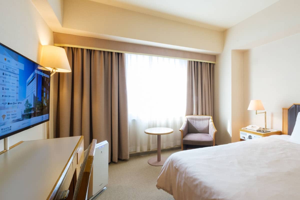 Une chambre d’hôtel comprenant un lit, un bureau, une chaise, une télévision, une fenêtre avec des rideaux et une table de chevet avec une lampe.