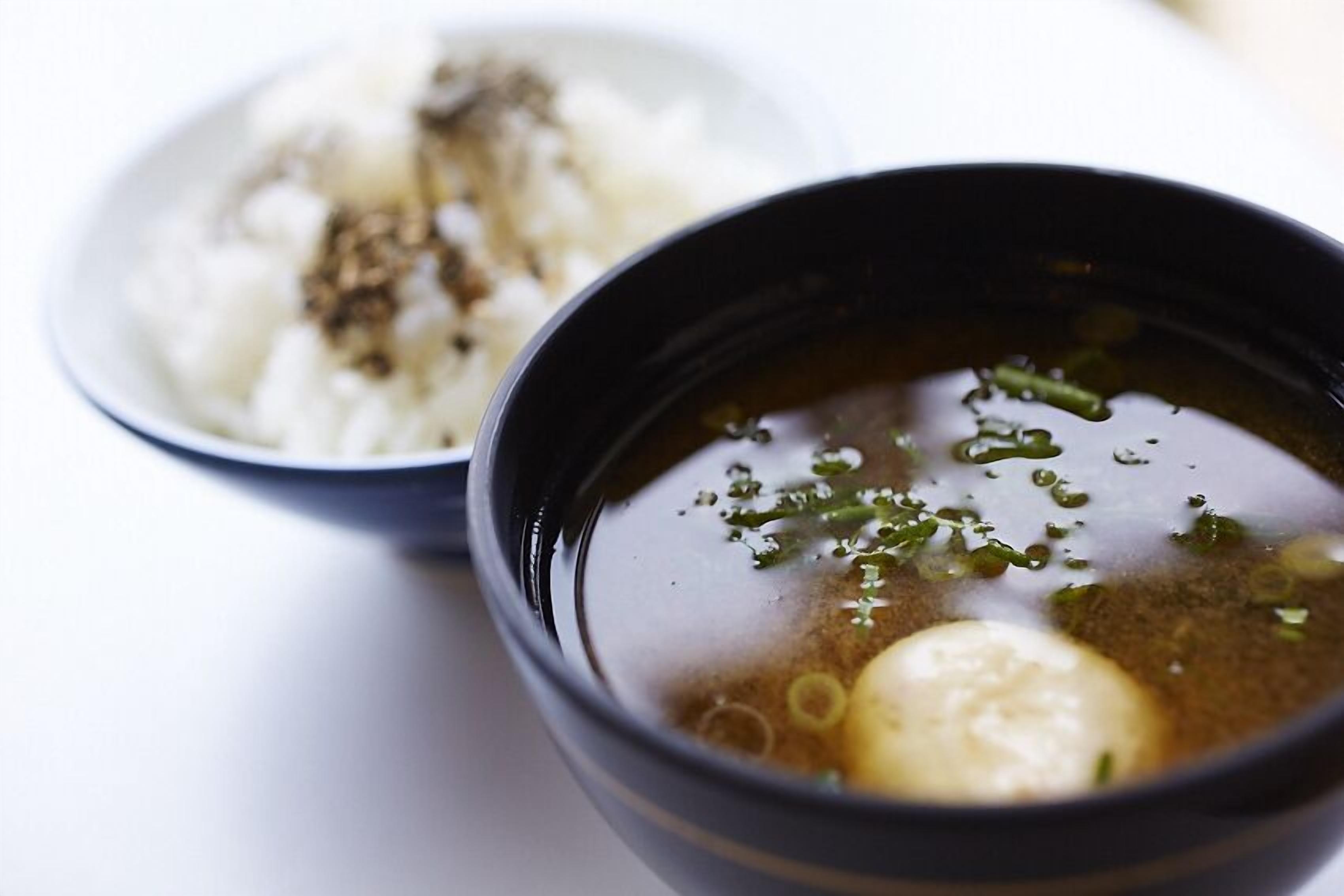 A bowl of clear soup with a boiled egg and green onions, accompanied by a bowl of white rice with brown seasoning.