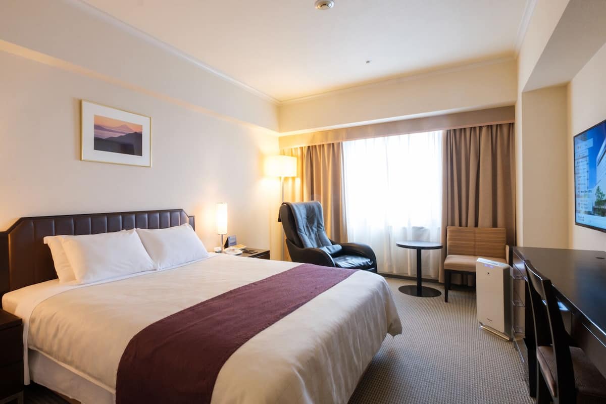 Une chambre d’hôtel avec un grand lit, un bureau, une chaise, une télévision et une fenêtre avec des rideaux.