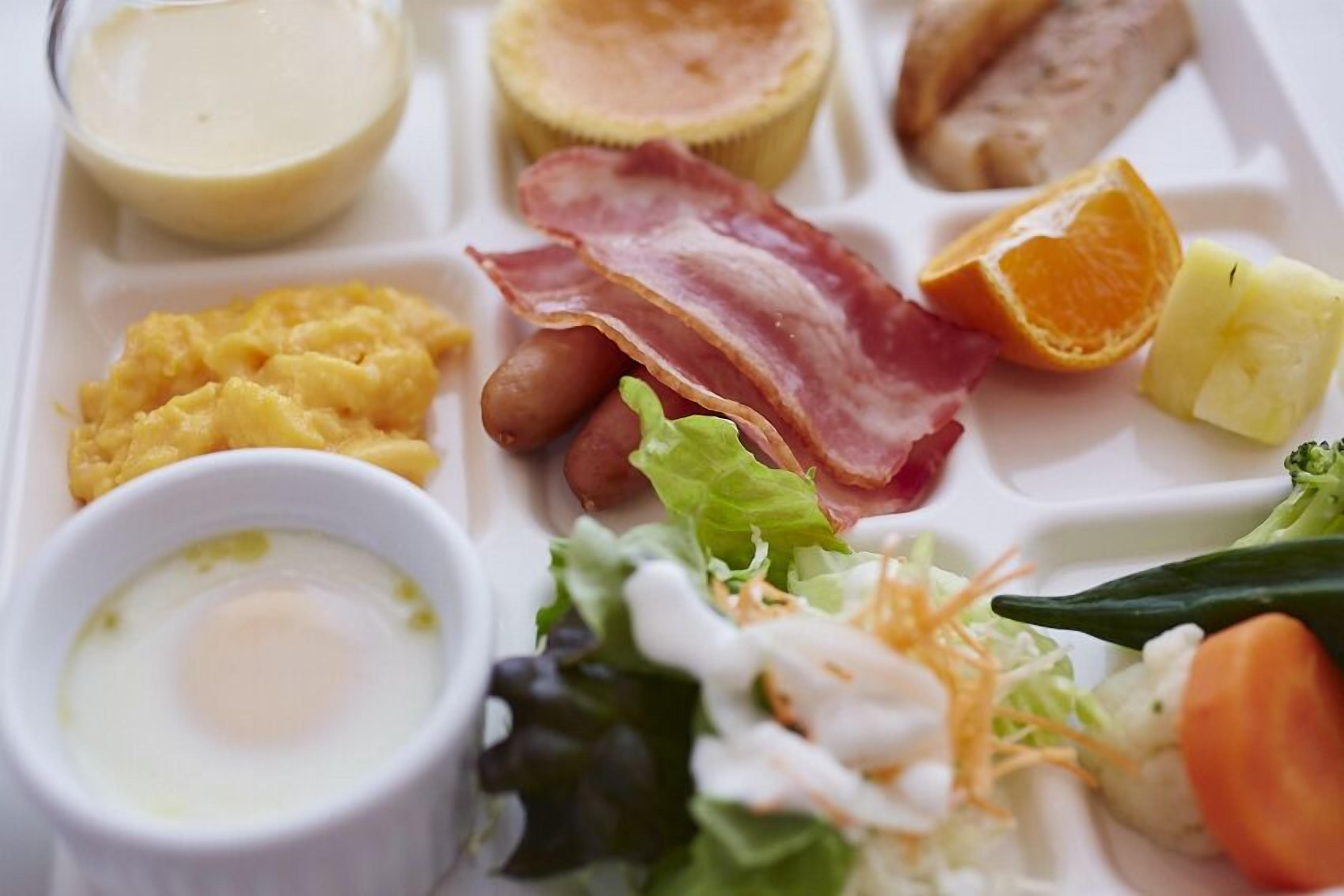 A meal tray with scrambled eggs, bacon, sausages, a slice of orange, a piece of pineapple, a small bowl of sauce, and a salad.
