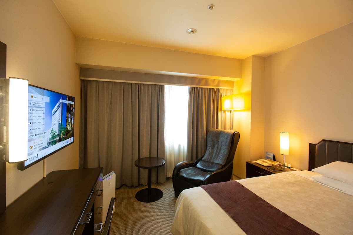 Une chambre d’hôtel comprenant un lit, un bureau avec un ordinateur, une chaise, une petite table, un téléviseur fixé au mur et une fenêtre avec des rideaux.