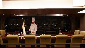 Un chef prépare un plat flambé à une table dans un restaurant offrant une vue sur la ville.