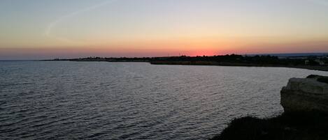 Solnedgang over kysten med rolige vand og en klippefyldt kystlinje.