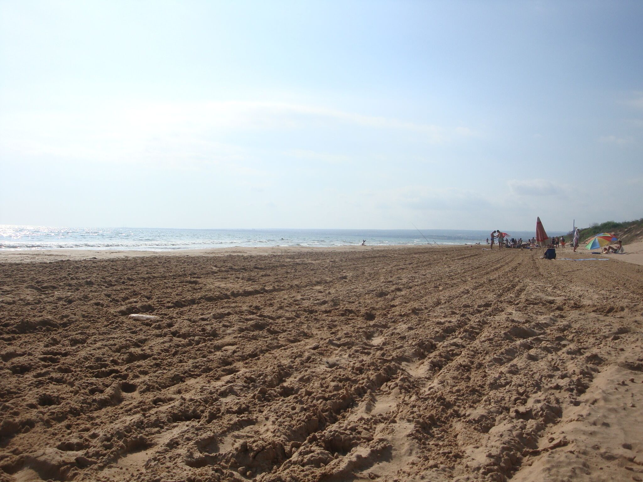 Ein weiter, sandiger Strand mit Fußabdrücken, die zum Wasser führen, einigen Sonnenschirmen und Menschen in der Ferne.