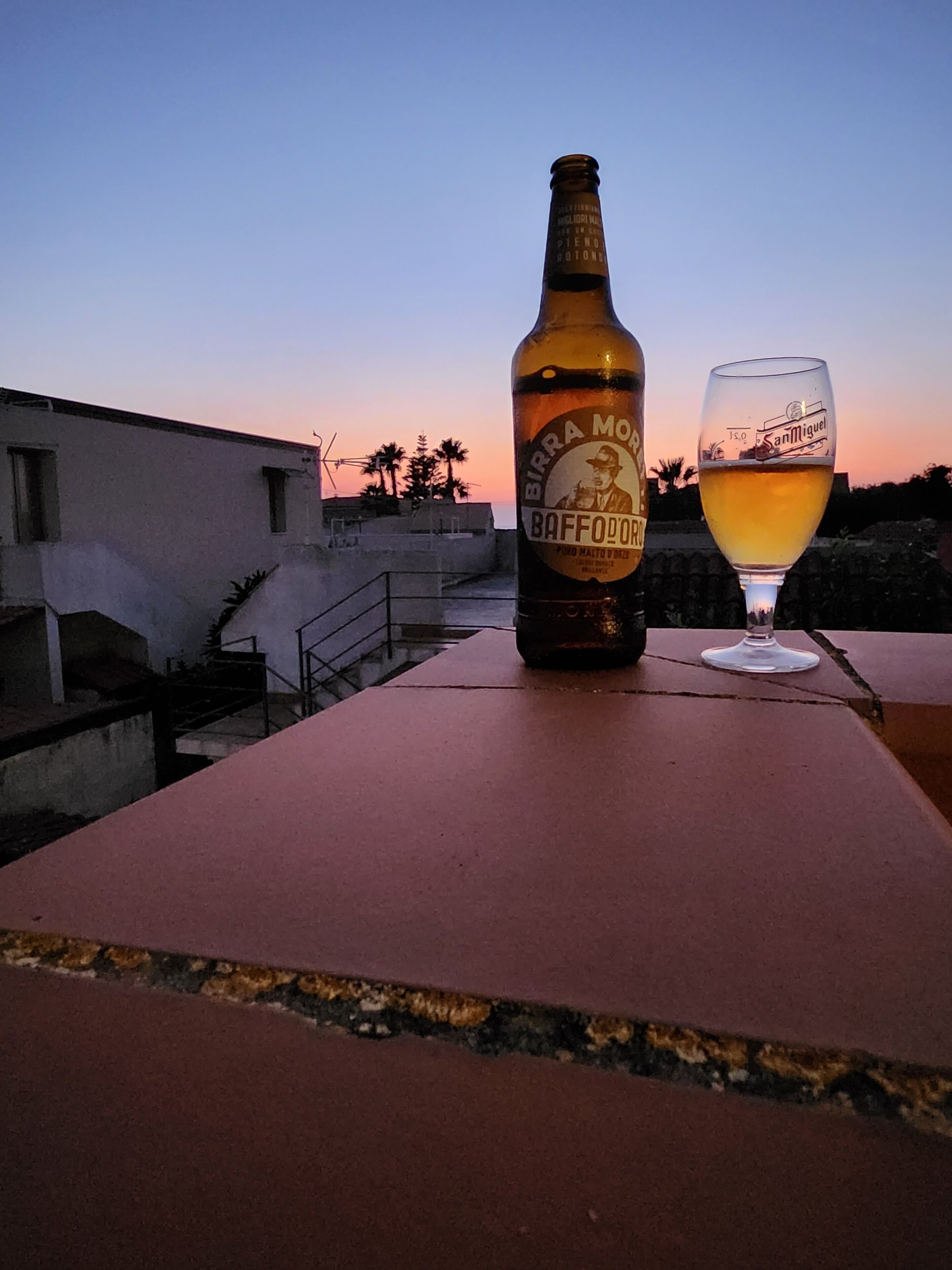 A bottle of Birra Moretti Baffo d'Oro beer and a glass on a table with a sunset view.