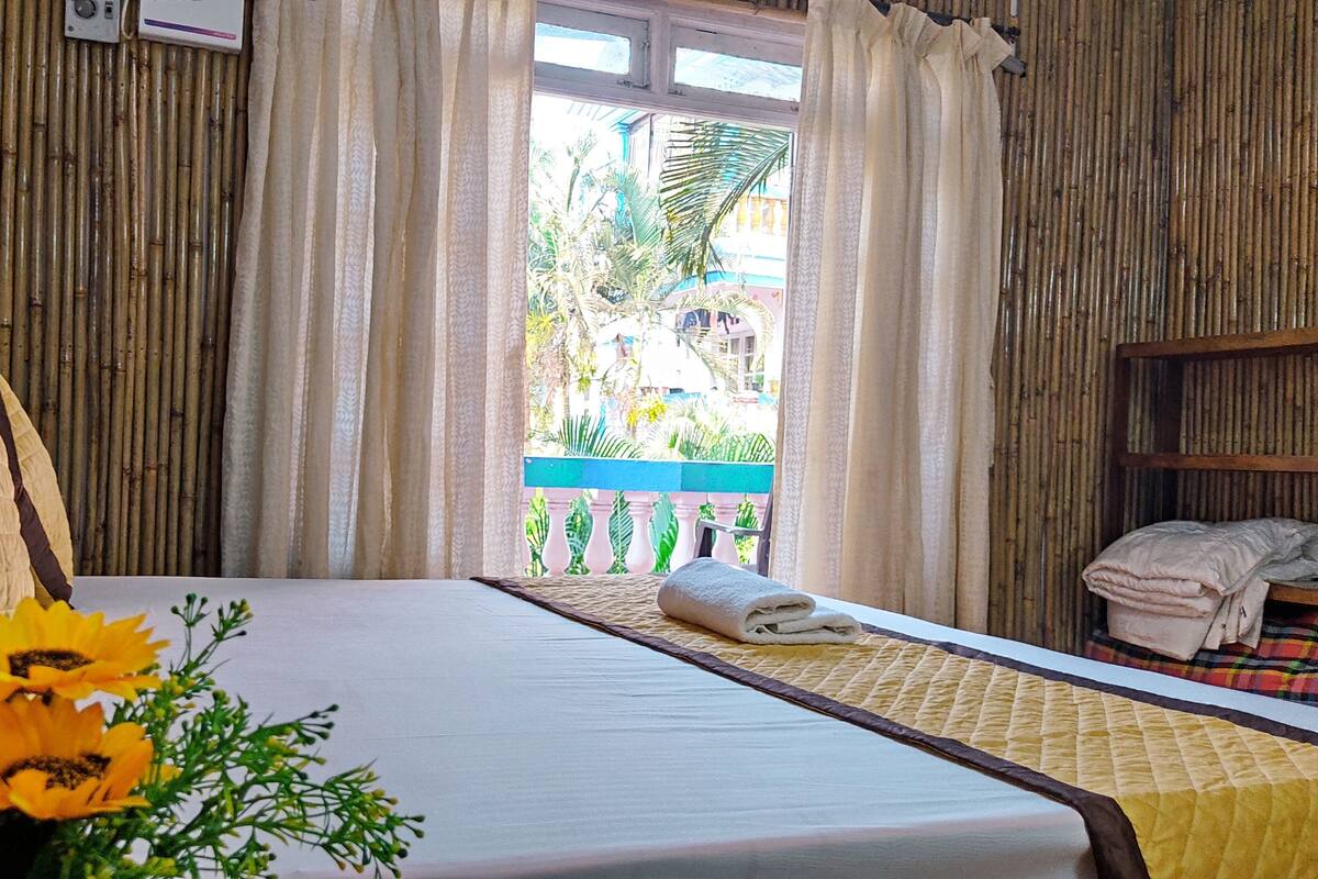 A room with a bed, a window with curtains, a view of greenery outside, and a small balcony with a blue railing.