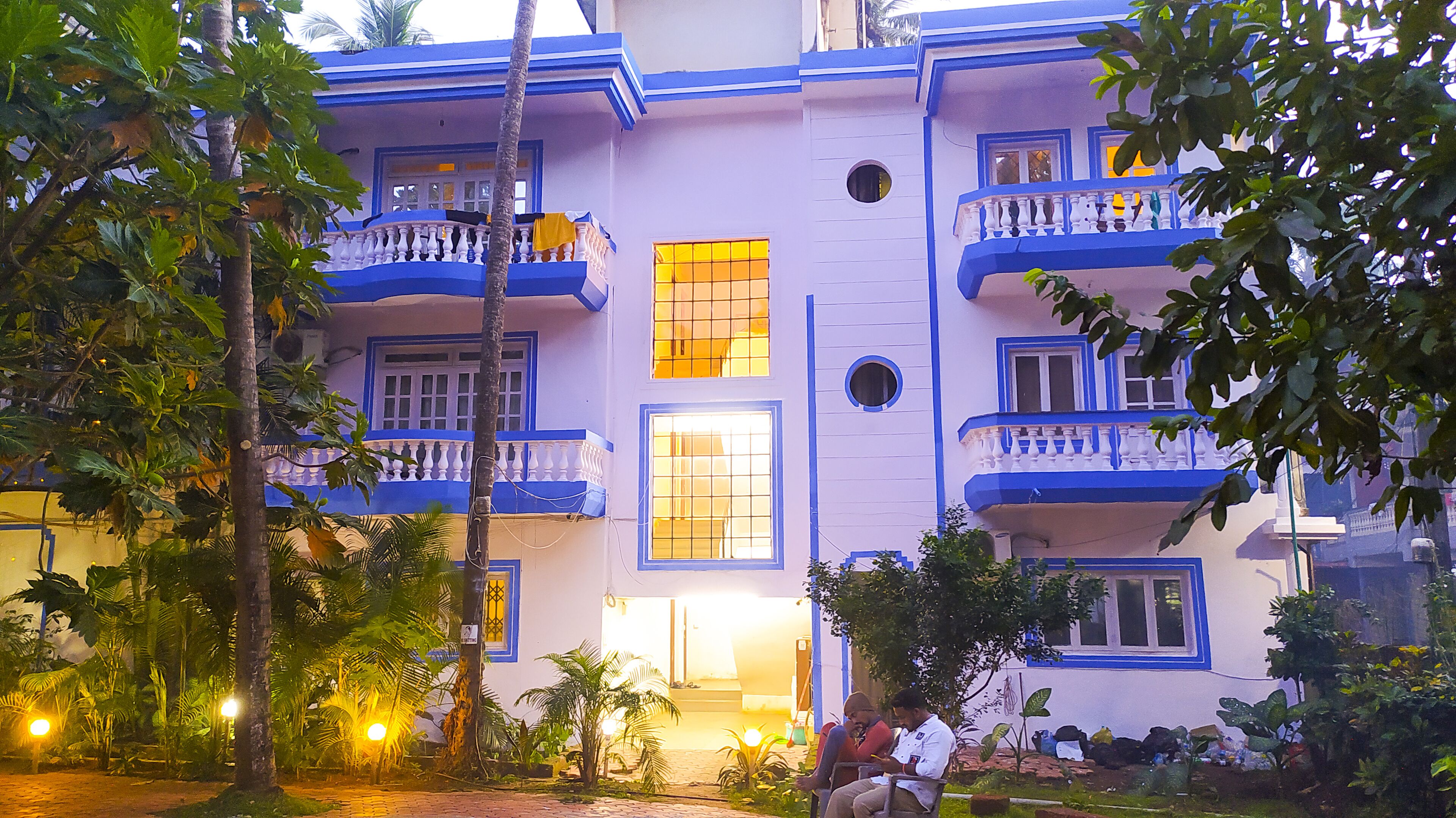 A multi-story building with balconies, large windows, and a garden area with trees and plants.