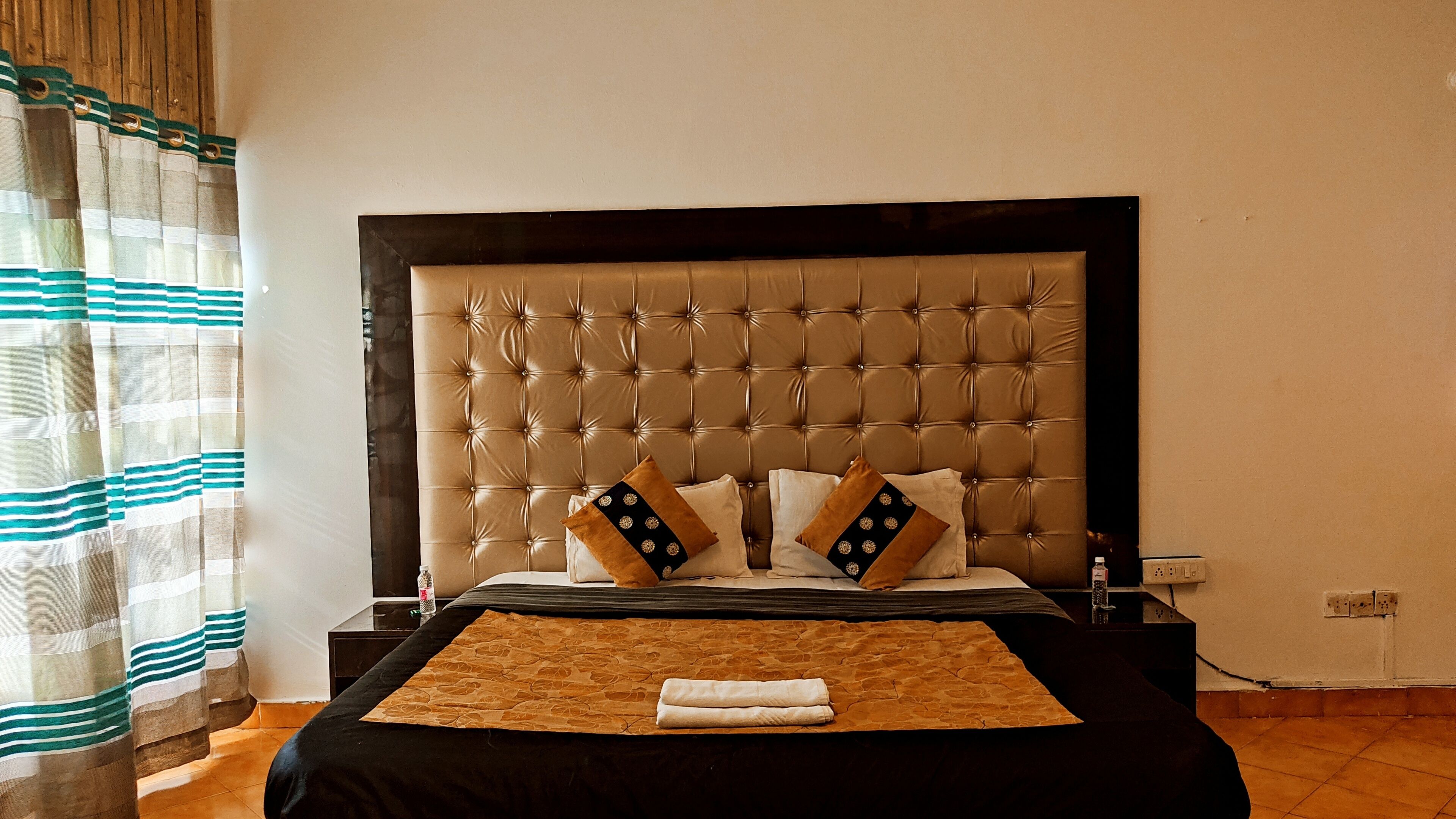 A hotel room with a large, tufted headboard, two pillows with decorative patterns, a neatly folded towel on the bed, and a striped curtain.