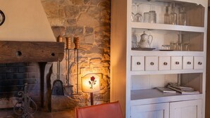 Ein rustikaler Raum mit einer Steinwand, einer Holzbank, einem Kamin mit traditionellem Design und einem weißen Schrank mit Glastüren, der Küchenutensilien beherbergt.