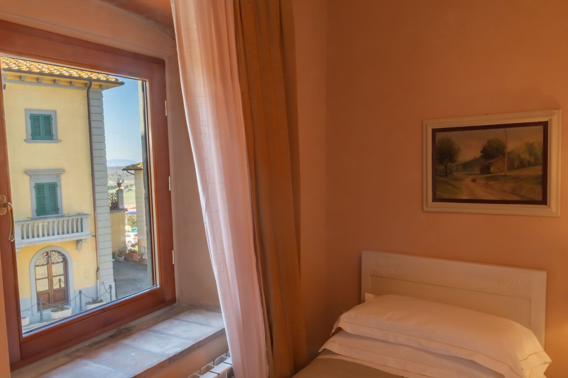 A bedroom with a bed, a window with a view of a building, and a painting on the wall.