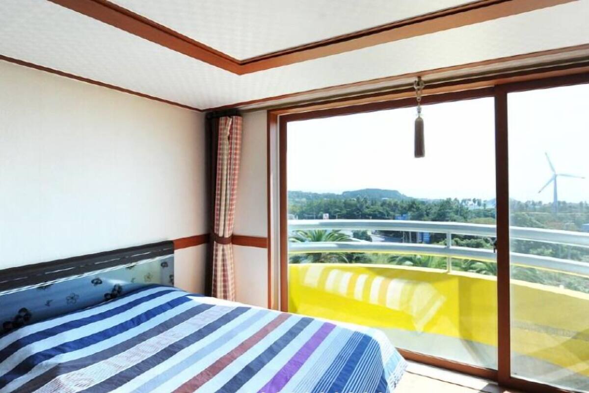 A bedroom with a large window, a bed with a striped bedspread, and a view of a green landscape with a wind turbine.