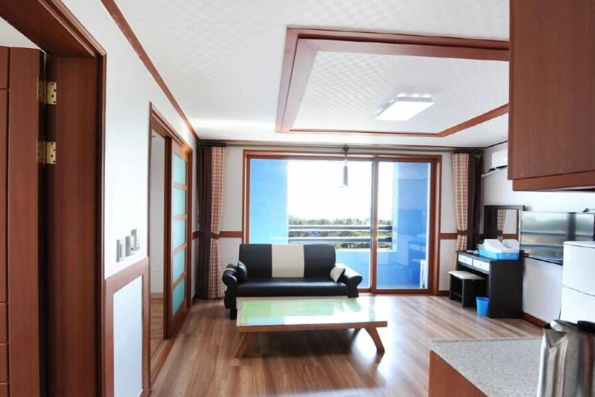 A compact living space with a sofa, a small table, a kitchenette, and a sliding glass door leading to a balcony.