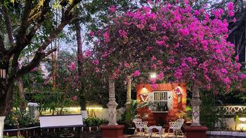 A garden patio with a stone floor, surrounded by lush greenery and pink bougainvillea flowers.