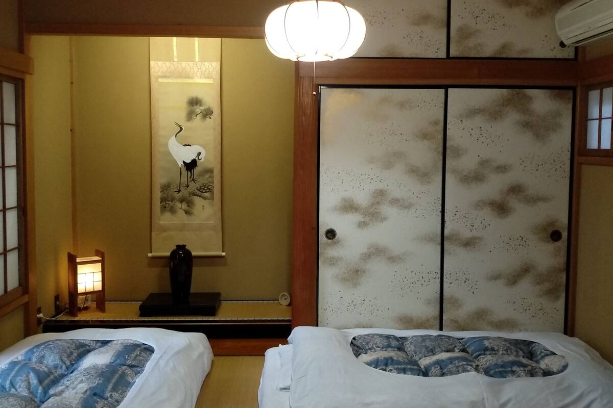 Ein traditioneller japanischer Raum mit Tatami-Boden, zwei Betten mit gemusterten Bettbezügen, einem Shoji-Bildschirm, einer Hängerrolle und einem Deckenventilator.