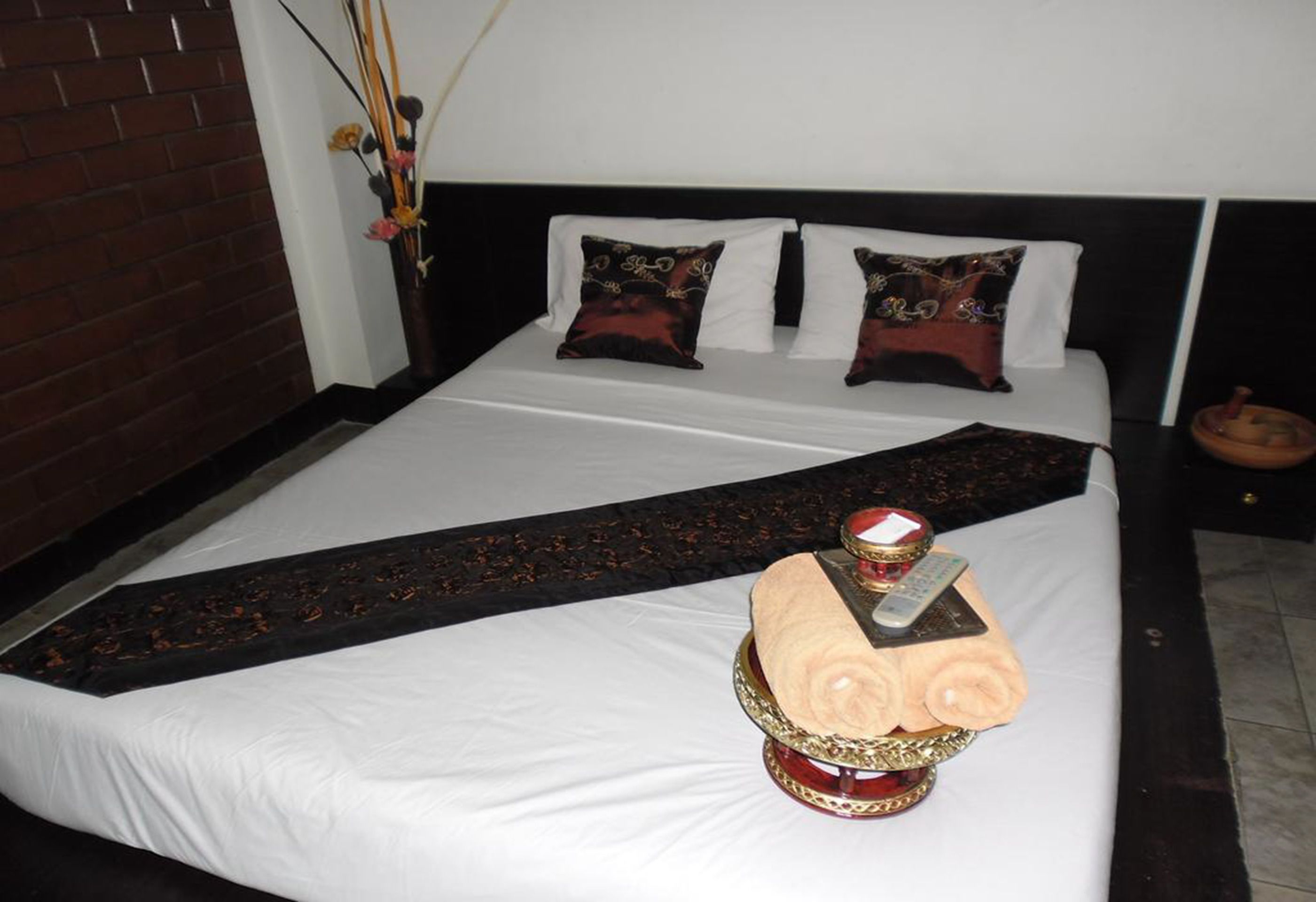 Una cama bien tendida con sábanas blancas, dos almohadones decorativos y un corredor negro. Un elemento central original es una escultura de madera que representa un rostro.