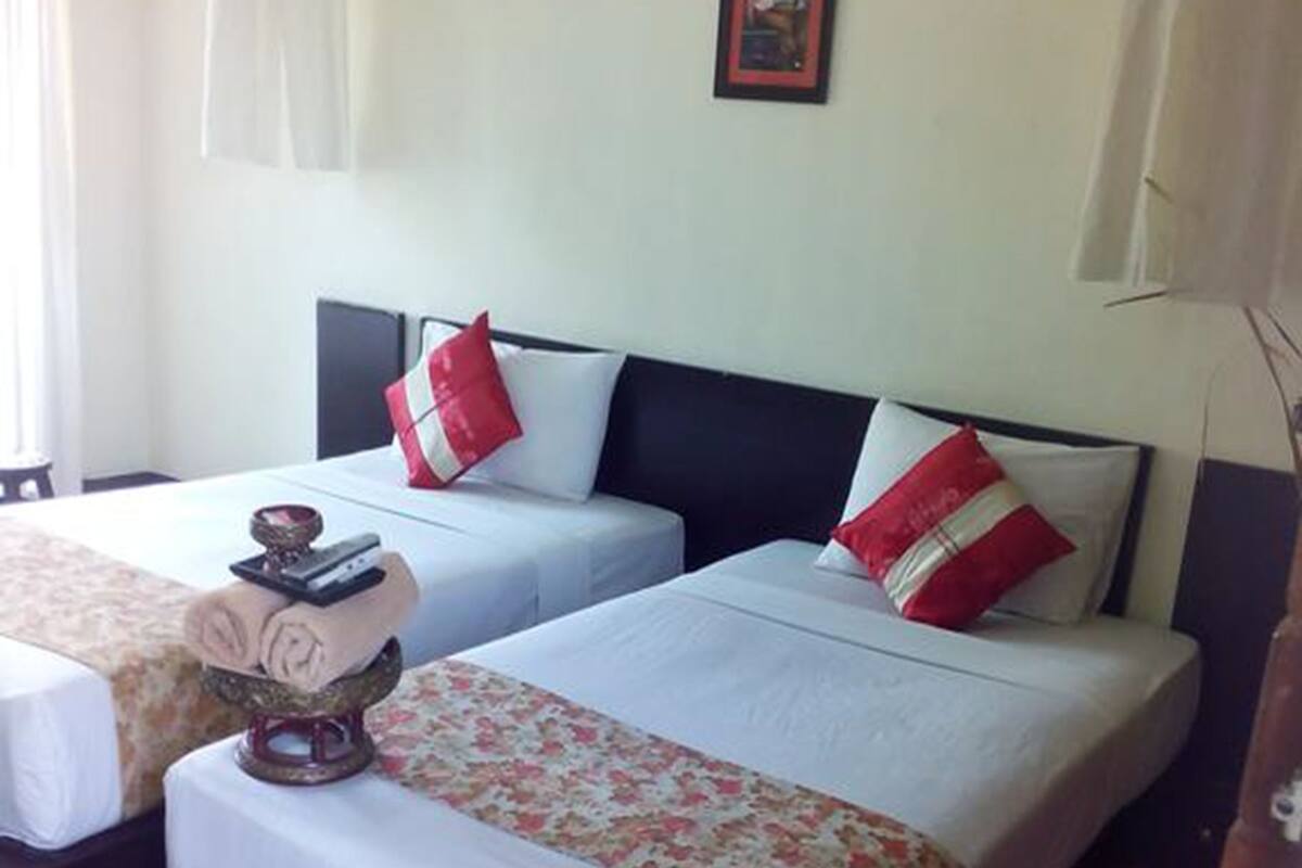 A hotel room with two beds, each with a white bedspread and red pillows. There is a framed picture on the wall and a hanging light fixture.