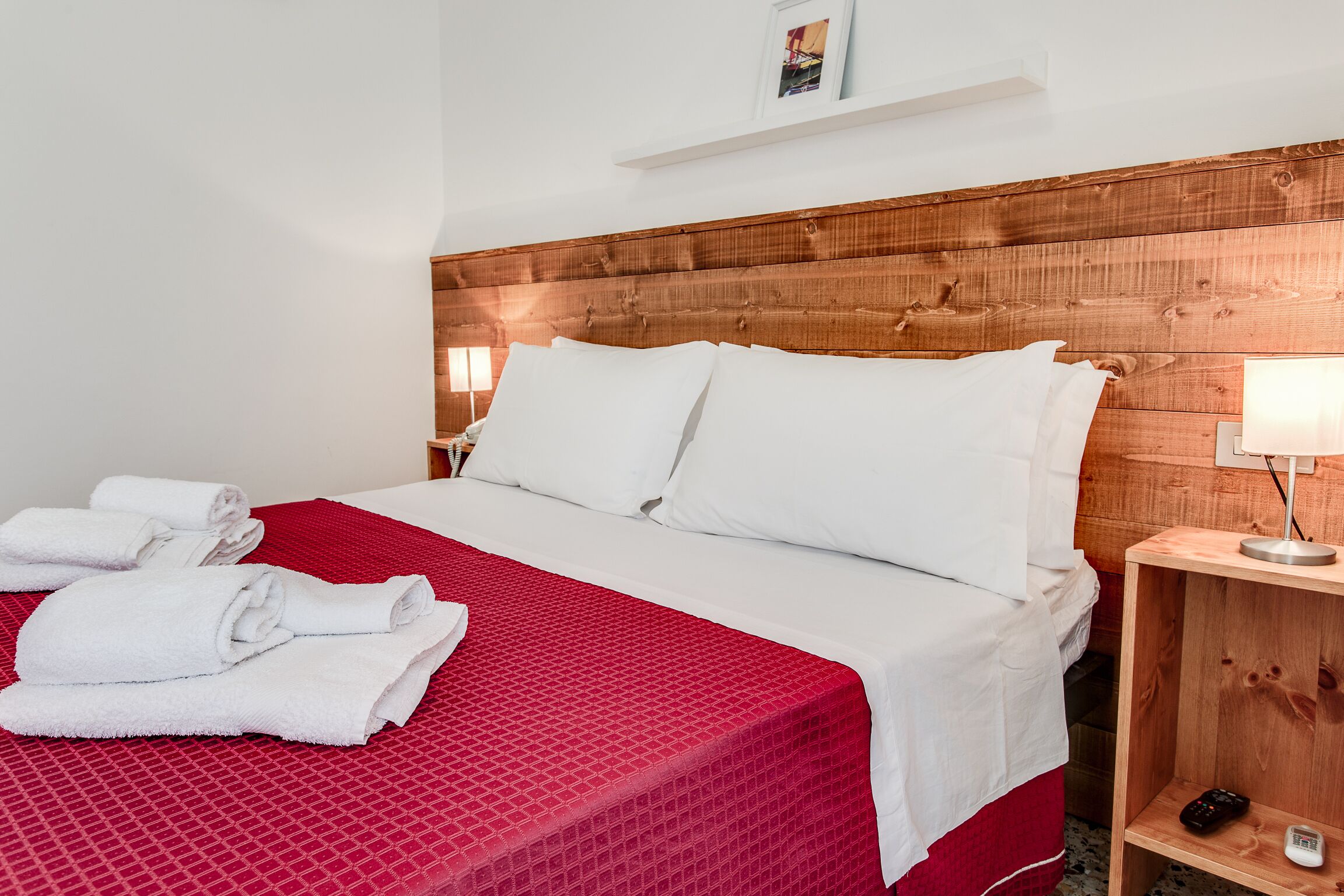 Una cama bien hecha con ropa de cama blanca y una colcha roja, un cabecero de madera, una mesita de noche con una lámpara, y un pequeño estante con una fotografía y un control remoto.