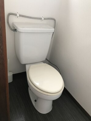 Een wit toilet met een beigekleurige zitting en deksel, een metalen pijp aan de muur bevestigd, en een donkere vloer.