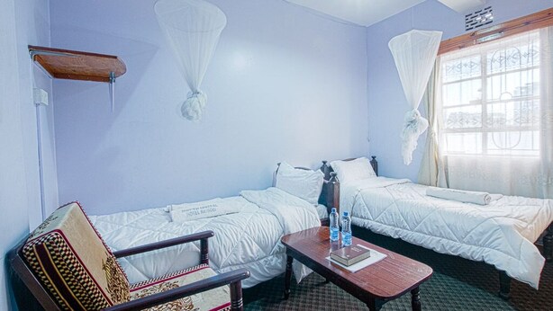 A room with two beds, a wooden chair, a small table, and a window with curtains. - Midview Hotel Nairobi (Nairobi)