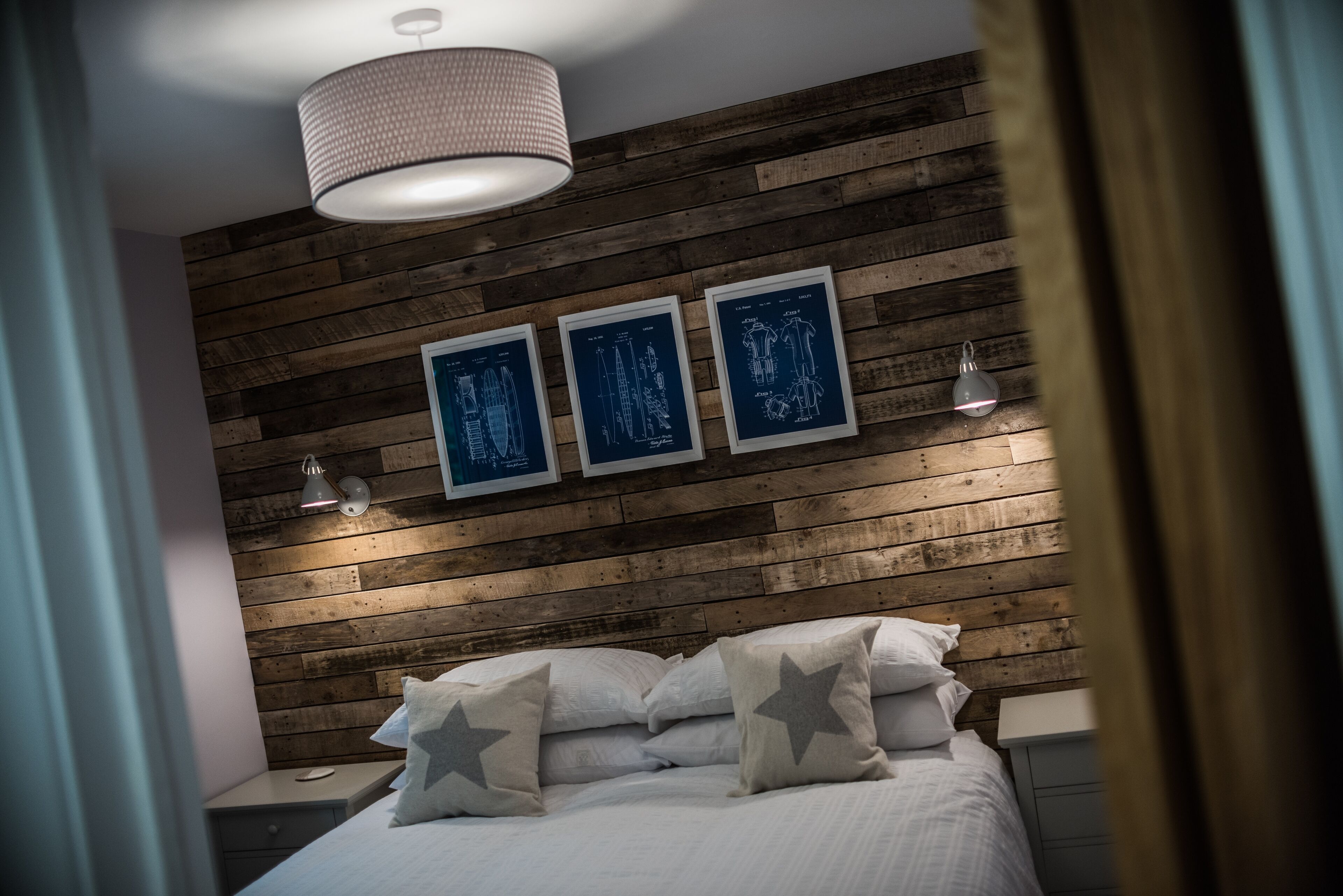 Une chambre avec une tête de lit en bois, un lit recouvert de linge de maison blanc et agrémenté d’oreillers à motifs étoilés, et trois dessins d’architecture encadrés, de couleur bleue, accrochés au mur.