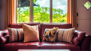 Un sofá de cuero rojo con cojines a rayas y de diseños geométricos, ubicado frente a una ventana con vistas a árboles verdes.