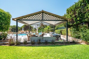 A covered outdoor seating area with a pool and greenery.