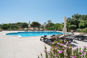 A swimming pool with lounge chairs and umbrellas, surrounded by greenery and a clear sky.