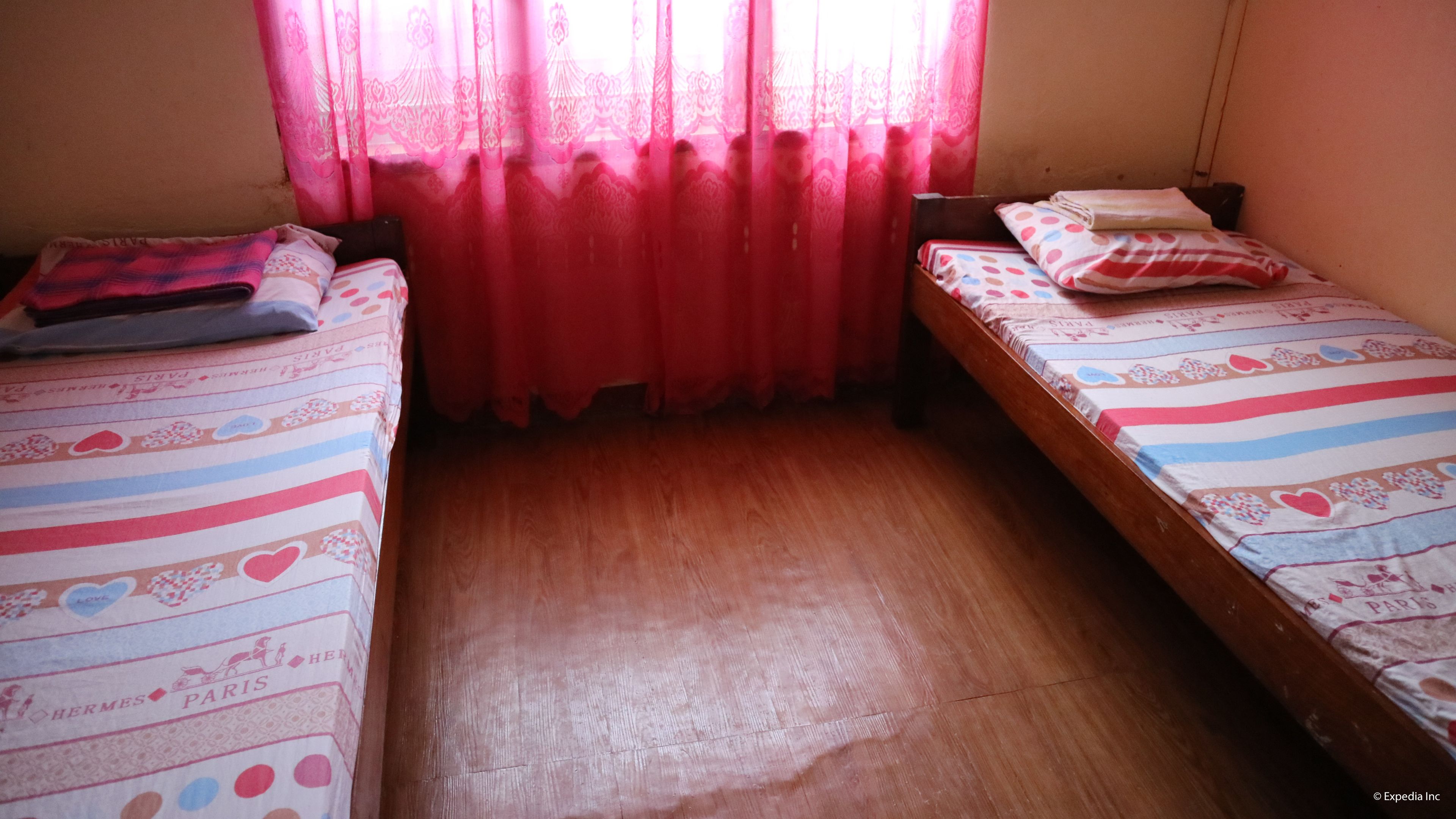 Een kleine kamer met twee eenpersoonsbedden, elk met een sprei met hartjesprint, een houten vloer en een raam met rode gordijnen.