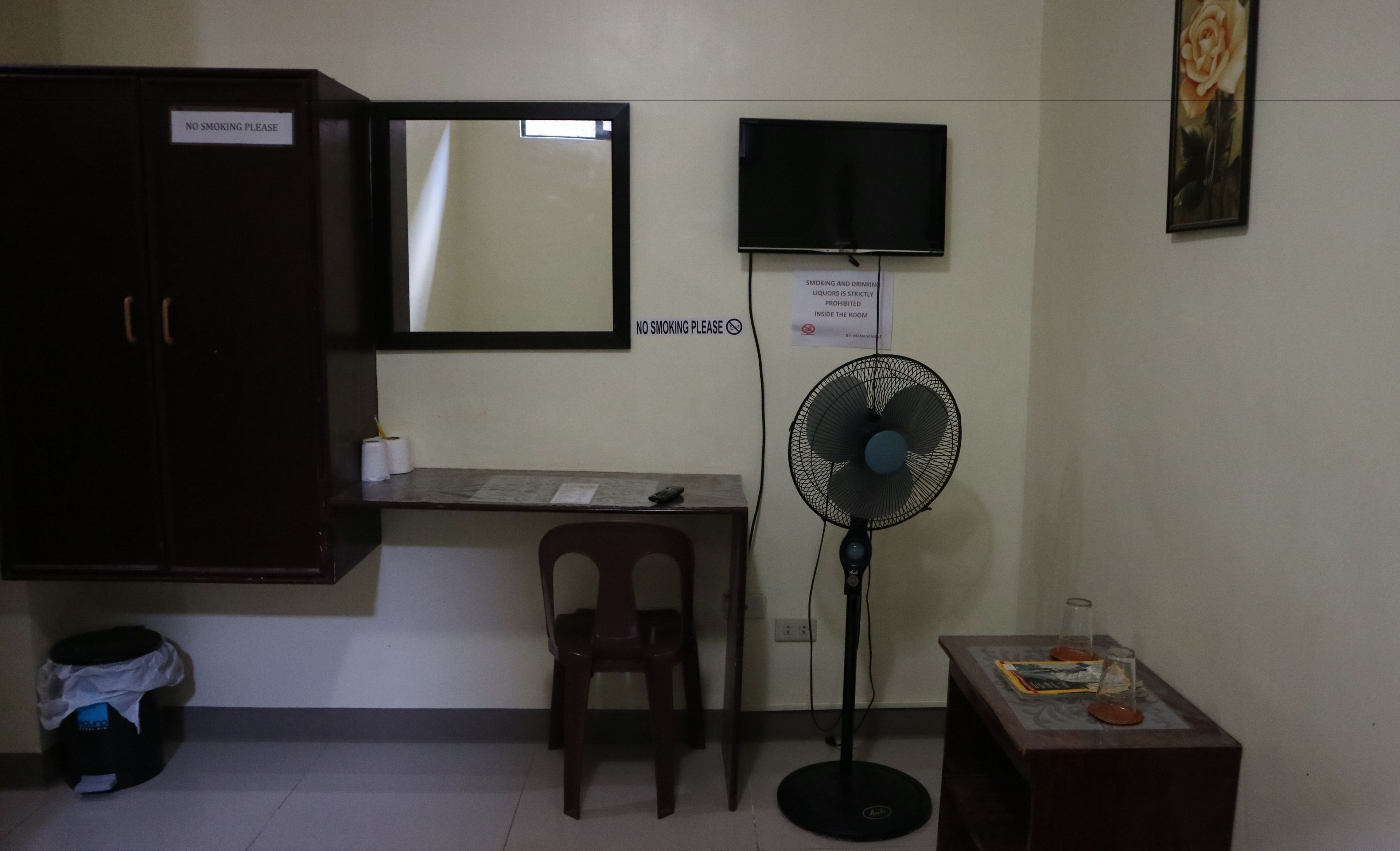 A small room with a desk, chair, and a standing fan. There are signs prohibiting smoking and drinking.