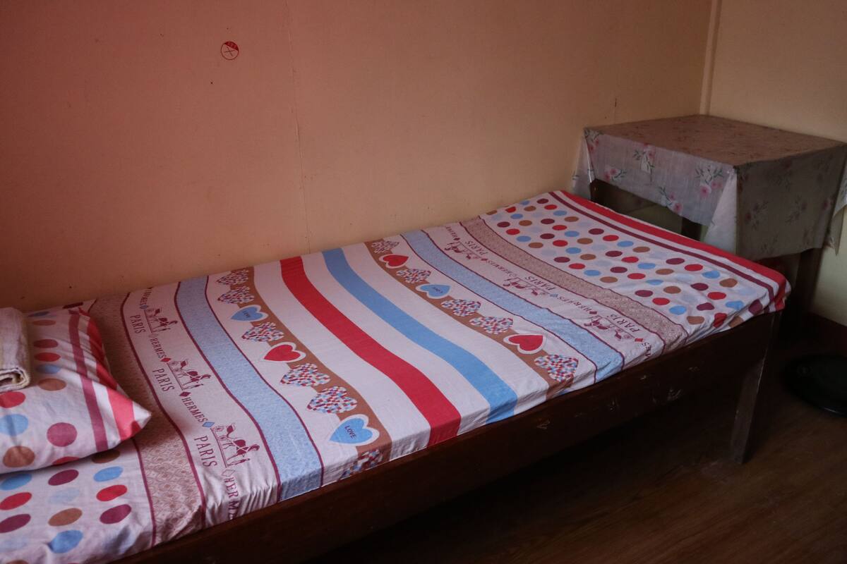 A small, single-bed room with a striped bedspread, pillows, and a small table with a floral tablecloth.