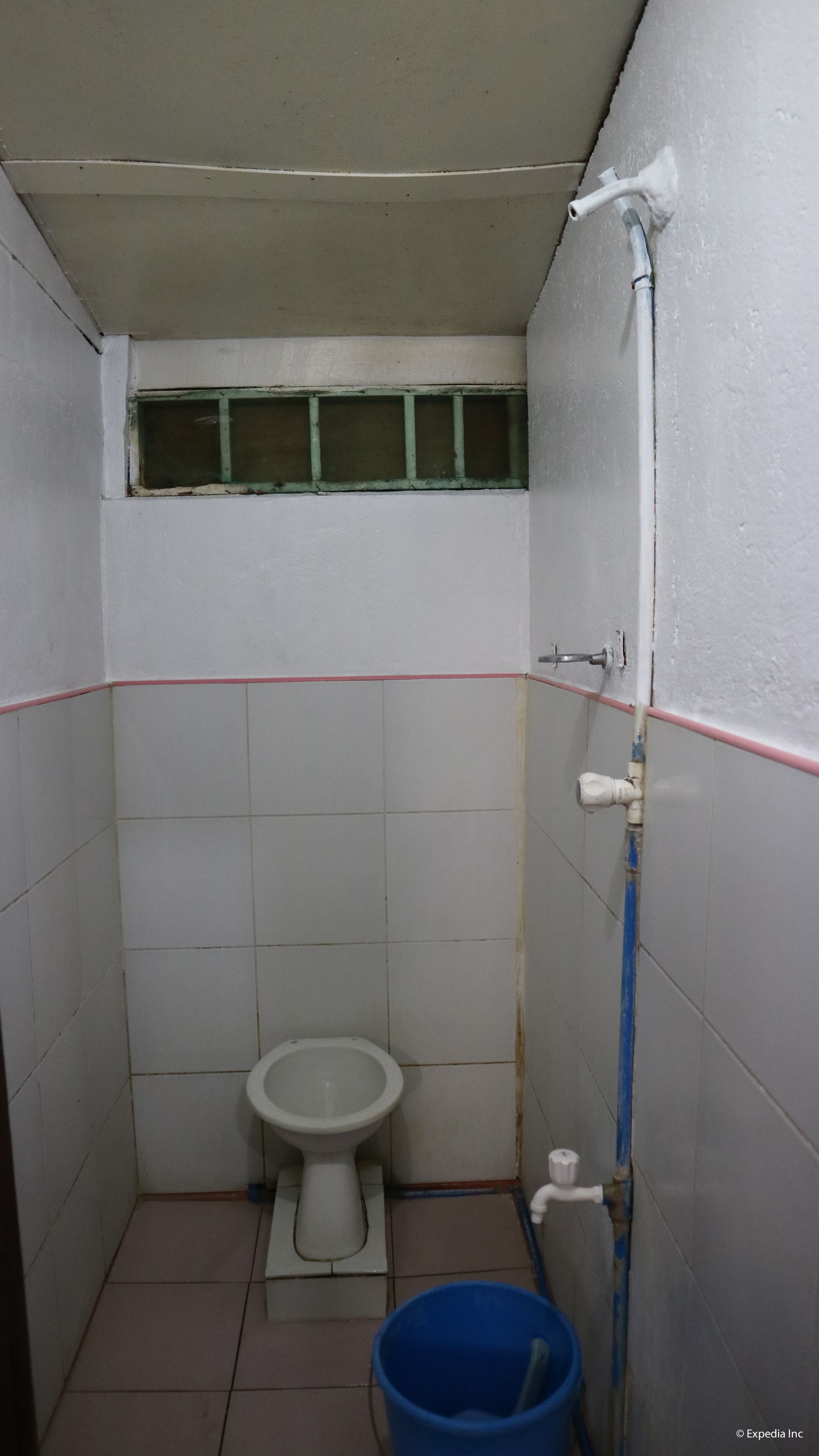 Une petite salle de bain simple, avec des toilettes, une fenêtre et un seau bleu.