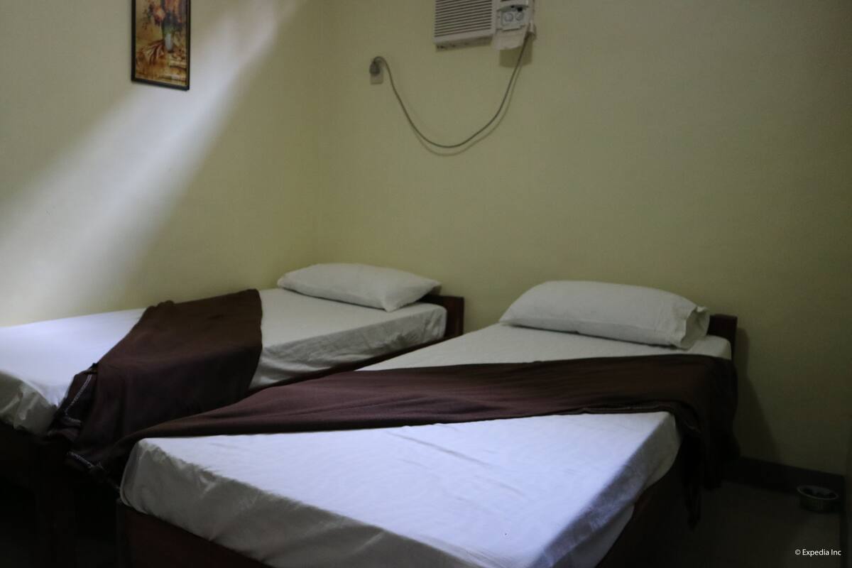 A small, dimly lit room with two single beds, each with white linens and a brown blanket. There is a wall-mounted air conditioning unit above the beds.