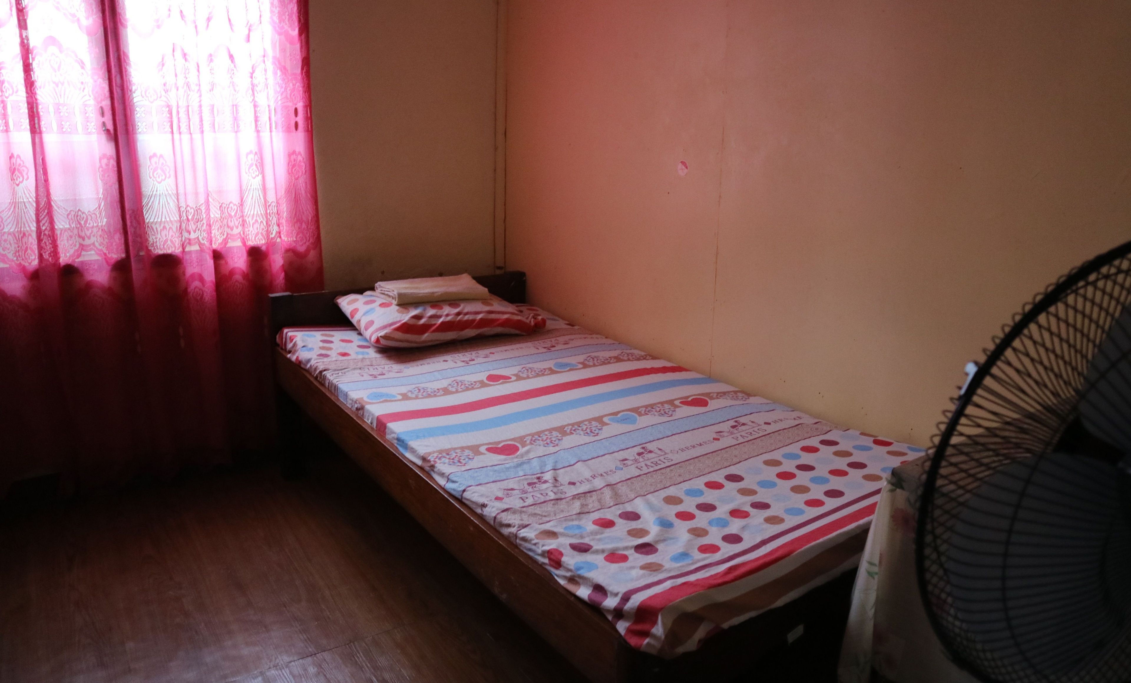 A small, single-bed room with a wooden floor, a bed with a striped bedspread, a pillow, and a standing fan.