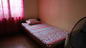 A small, single-bed room with a wooden floor, a bed with a striped bedspread, a pillow, and a standing fan.