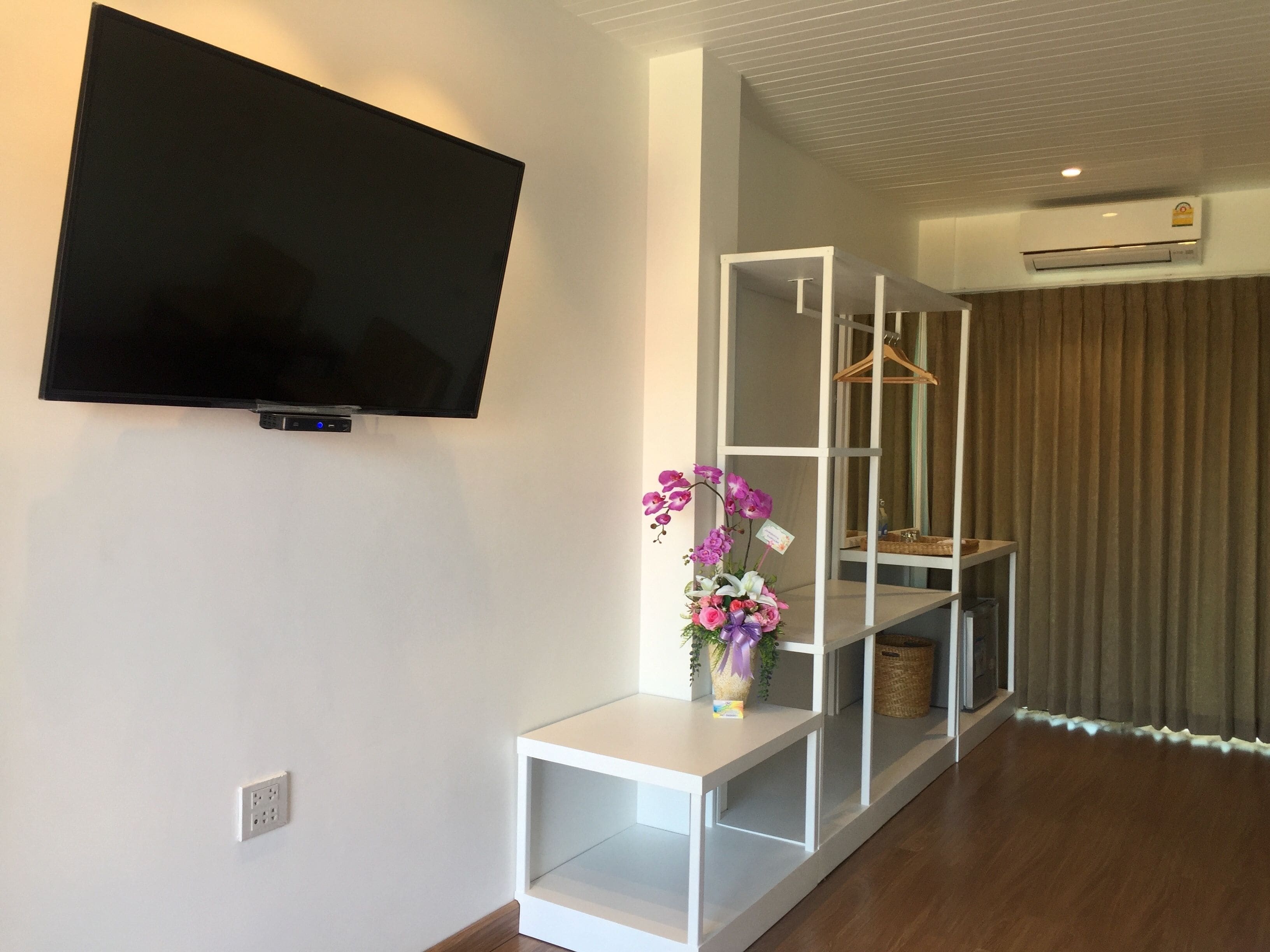 Una sala de estar moderna con un televisor montado, estanterías blancas, una mesita con un arreglo floral y una cortina marrón.