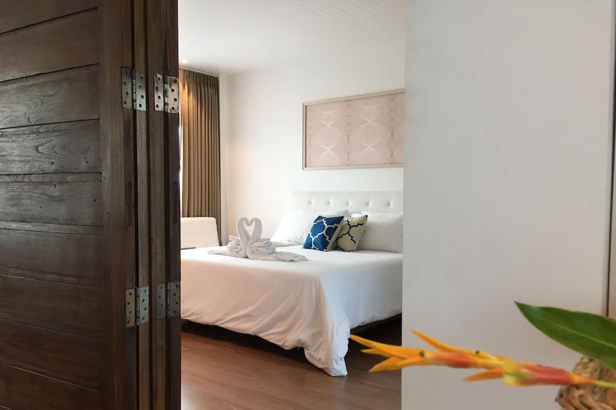 Une chambre d’hôtel avec une porte en bois, un lit avec une literie blanche, un oreiller bleu et blanc, et un luminaire fixé au mur.