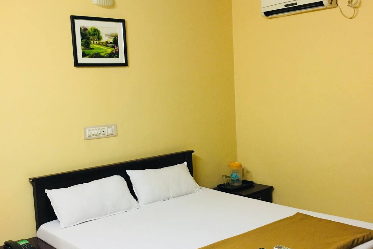 A hotel room with a bed, a nightstand, a bedside table, a ceiling fan, an air conditioning unit, and a framed picture on the wall.