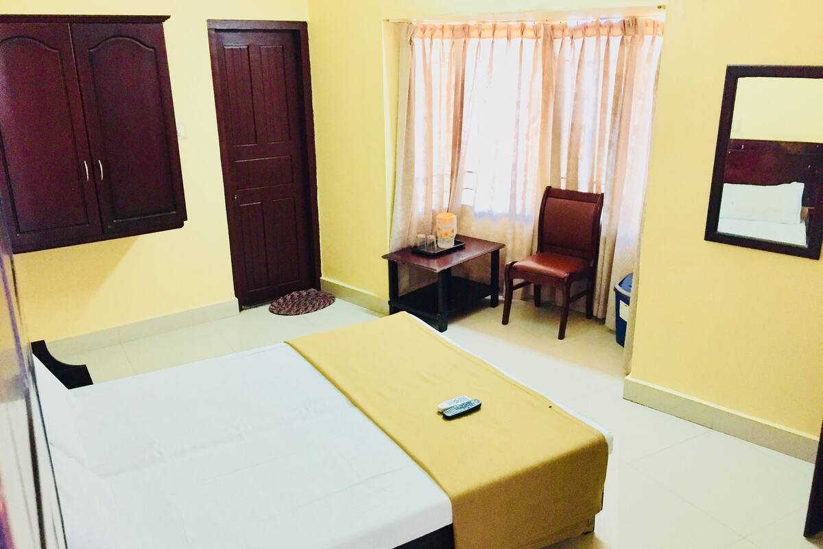 A hotel room with a bed, a chair, a small table with a lamp, and a window with curtains.