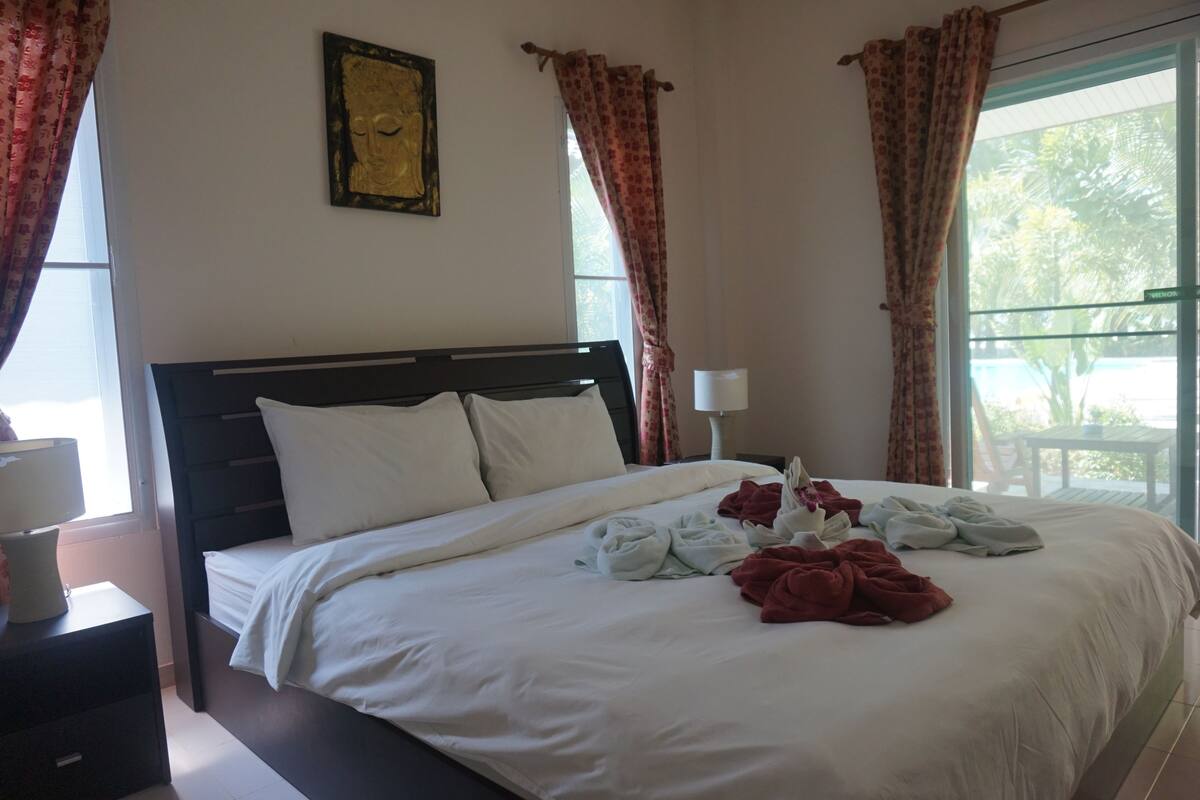 A neatly made bed with white linens and a red blanket, a bedside table with a lamp, and a window with curtains.