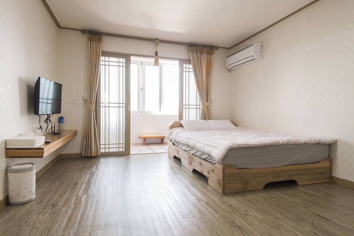 Une chambre à coucher avec un lit en bois, un téléviseur posé sur une étagère, une poubelle et un appareil de climatisation.