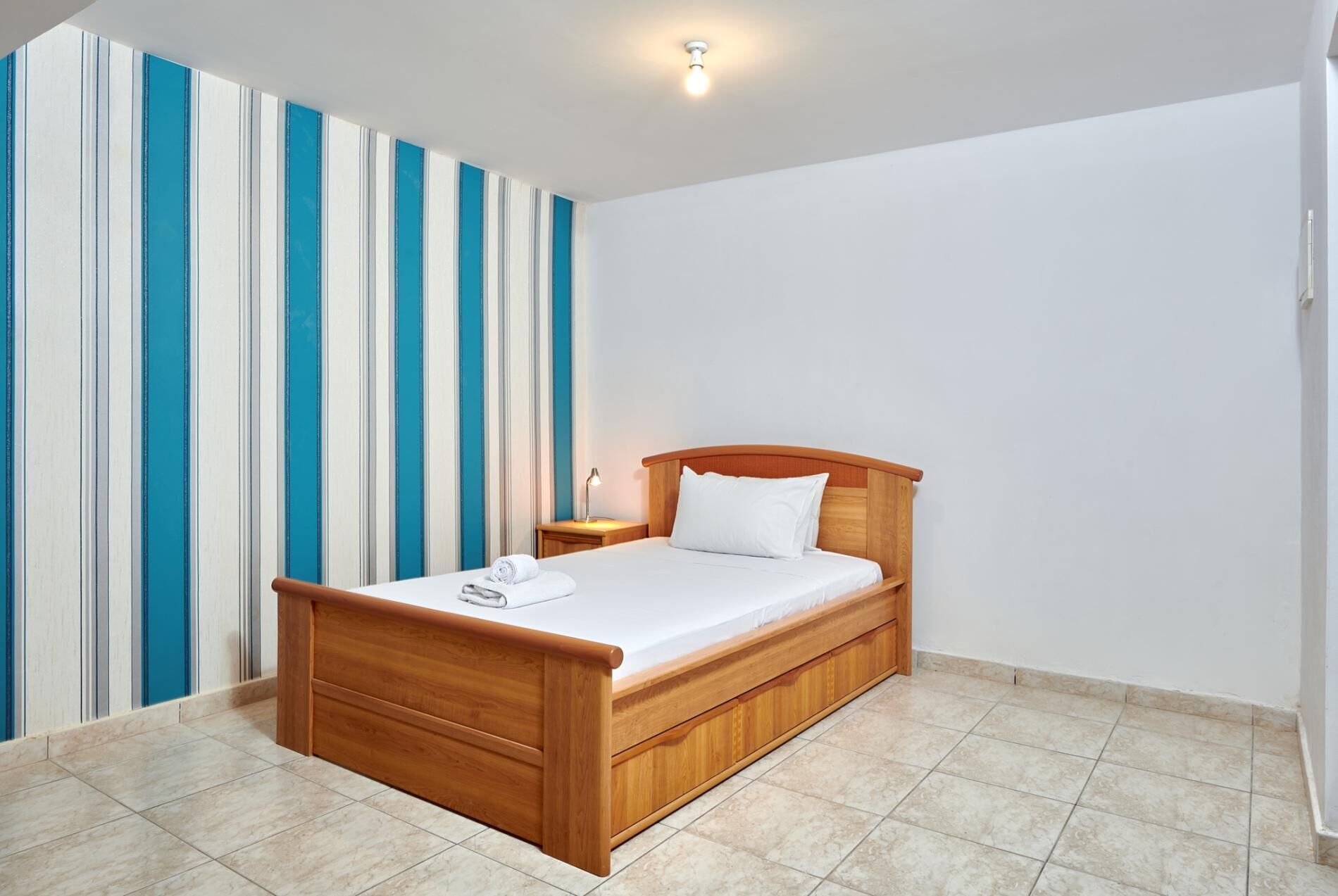 Una cama individual con cabecera de madera en una habitación con papel tapiz a rayas y suelo de baldosas.