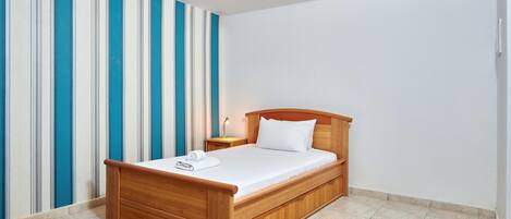 Una cama individual con cabecera de madera en una habitación con papel tapiz a rayas y suelo de baldosas.