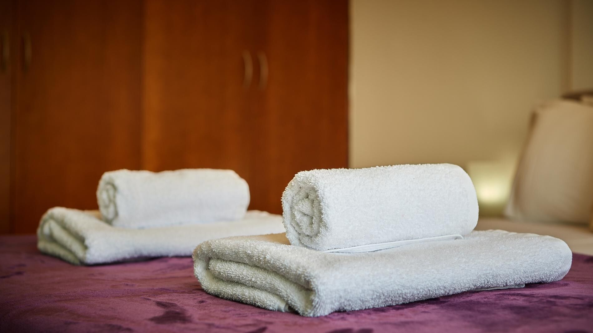 Dos toallas blancas enrolladas sobre un edredón morado en una habitación con muebles de madera.
