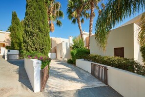 A villa with a paved driveway, surrounded by palm trees and a white wall with a gate.