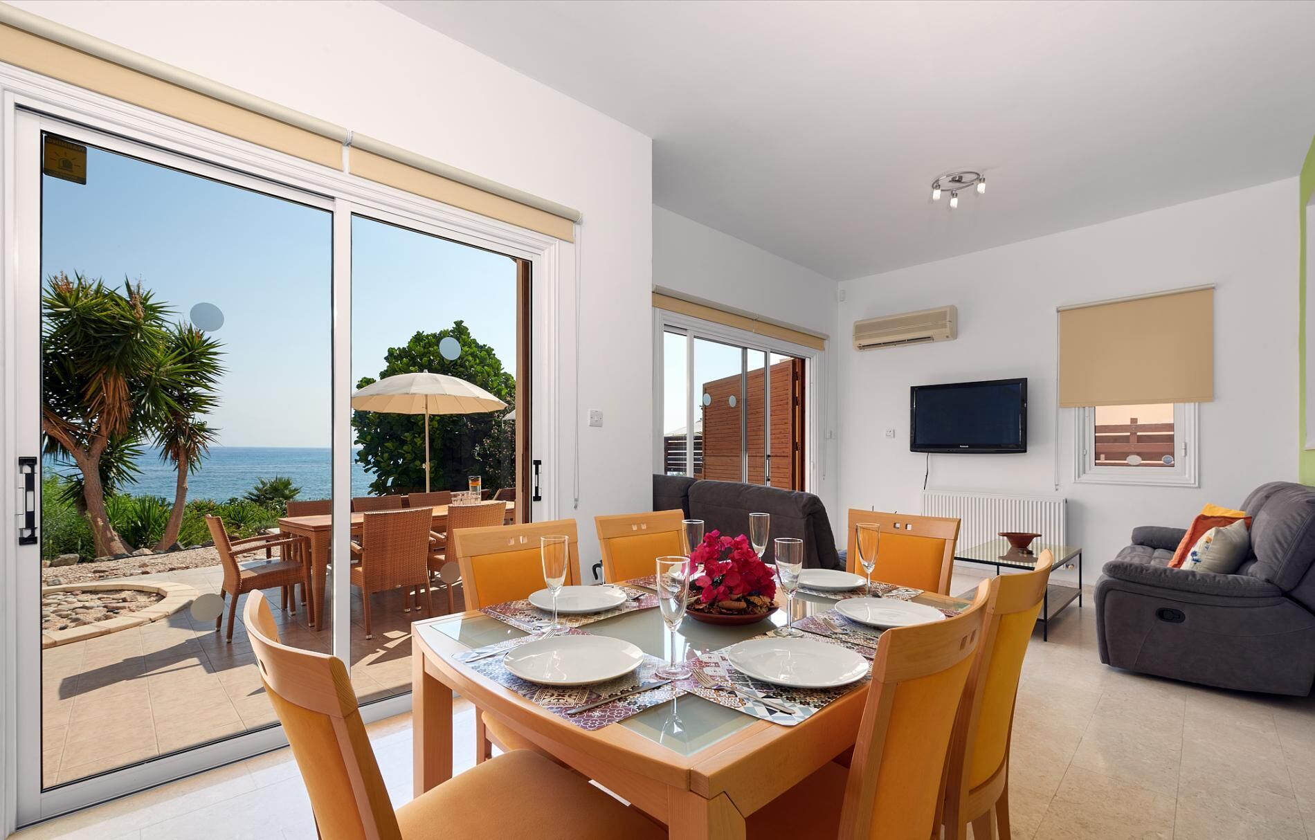 A dining area with a table set for four, a sofa, a TV, and a view of the sea.