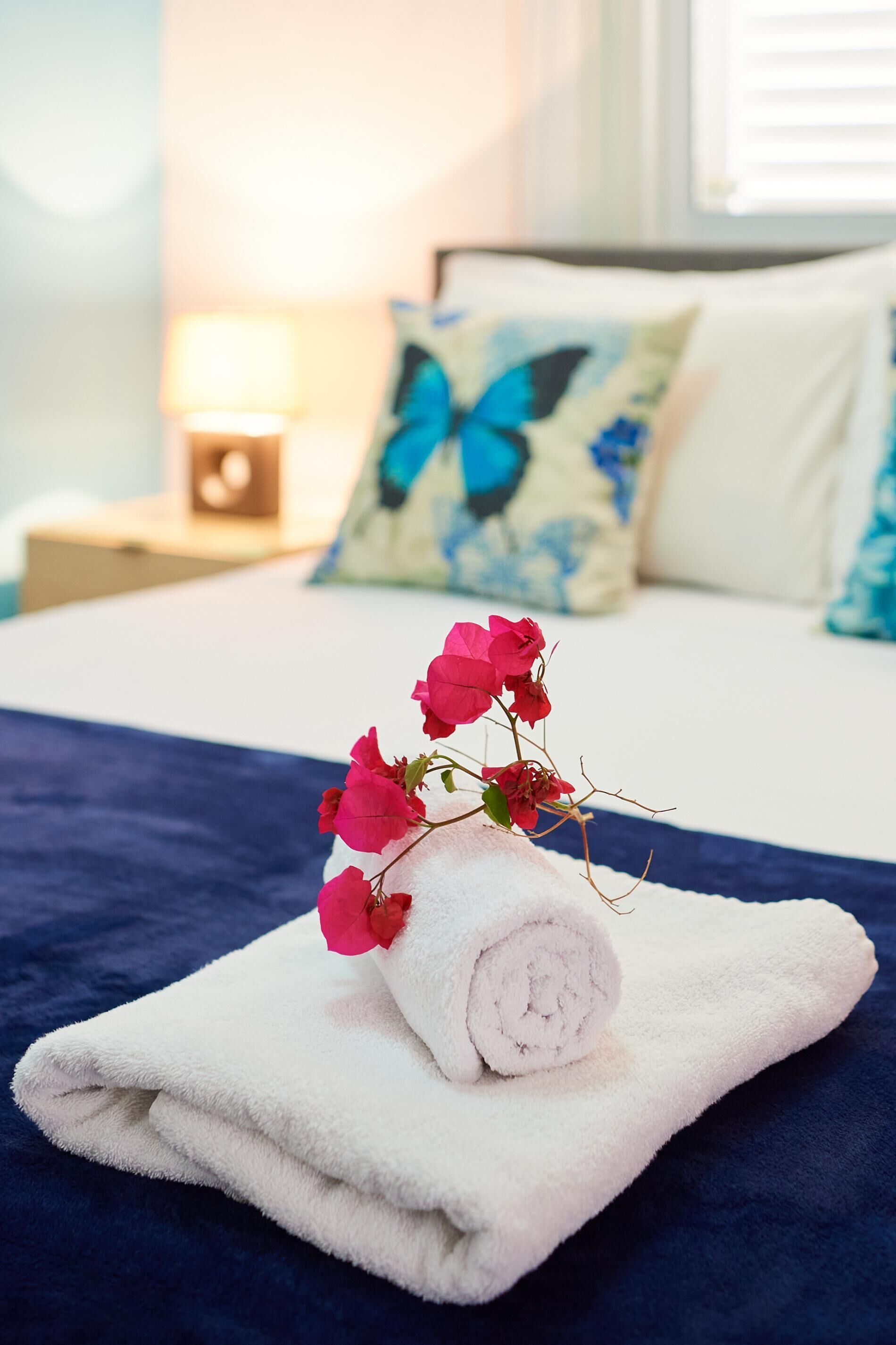 A neatly made bed with a rolled white towel and pink flowers on a blue bedspread.
