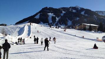 En skidbacke med skidåkare och snowboardåkare, en stuga och en bergskuliss i bakgrunden.