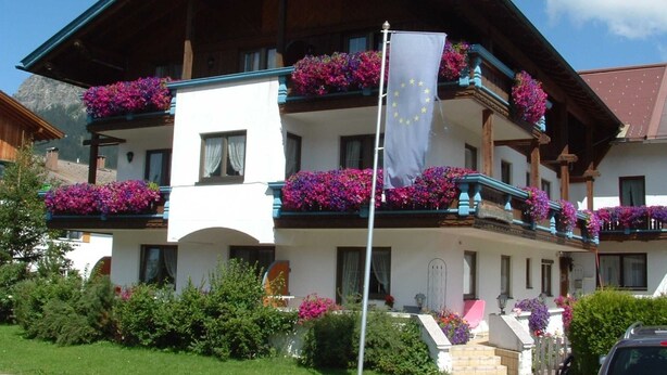A two-story building with balconies adorned with flowers, a flagpole with a flag, and a clear sky.