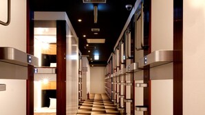 Un pasillo de hotel con un diseño de piso a cuadros, luminarias modernas y una escalera central que lleva a una habitación con cama y mesita de noche.